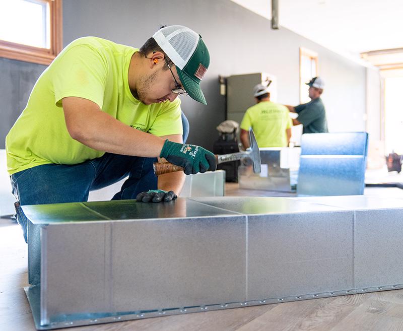 McElroy’s residential heating and air technician, Zach Brinker, assembles ductwork while PJ Rocha and Ben Walker install a furnace in an outbuilding that’s transforming into a space for exercise and workouts.